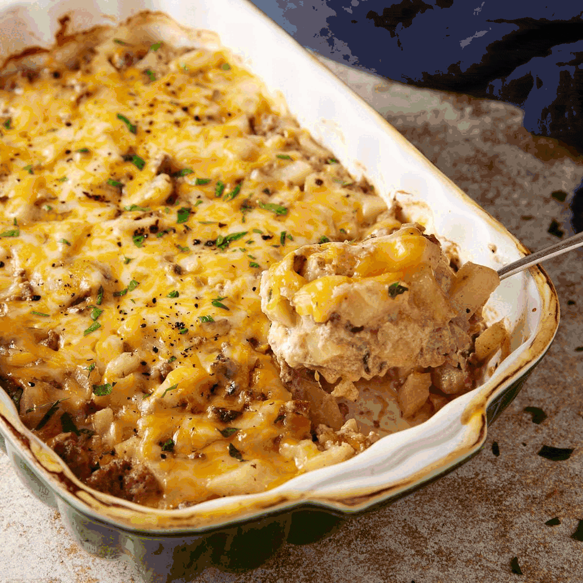 A casserole dish filled with a cheesy, baked ground beef and potato casserole. A serving spoon is lifting out a portion, showing layers of cheese, beef, and potatoes, topped with fresh herbs.