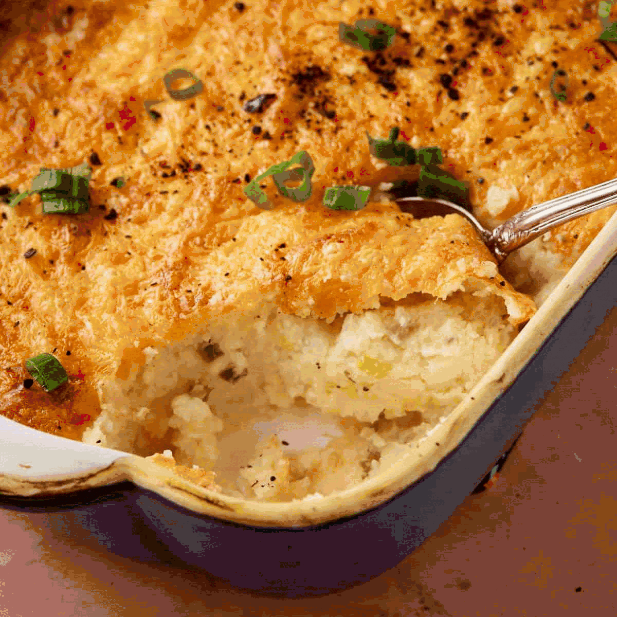 A casserole dish filled with golden-brown baked mashed potatoes, topped with melted cheese and chopped green onions. A serving spoon reveals the creamy potato filling inside.