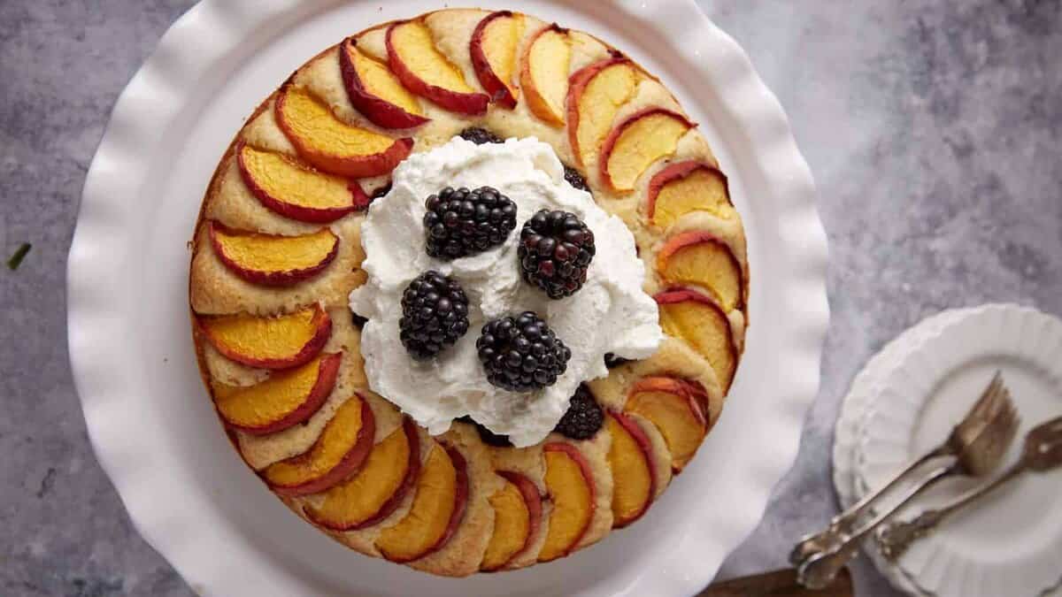A round cake topped with peach slices arranged in a circle, a large dollop of whipped cream, and several blackberries in the center, served on a white plate with dessert plates and utensils beside it.