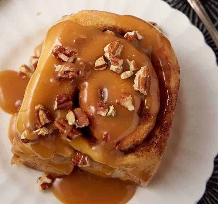 A cinnamon roll topped with caramel sauce and chopped pecans sits on a white scalloped plate, offering one of the coziest desserts for National Hot Mulled Cider Day. A silver fork rests beside it on a dark patterned surface.