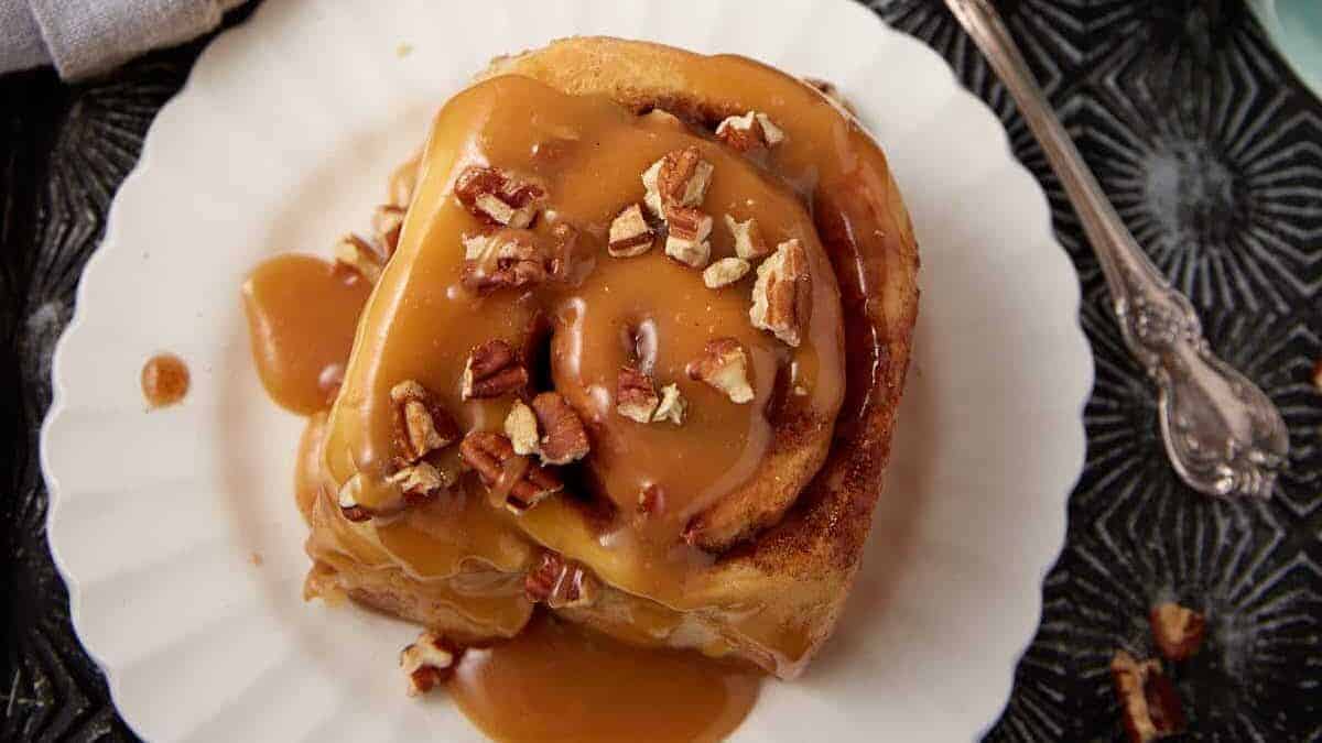 A cinnamon roll topped with caramel sauce and chopped pecans sits on a white scalloped plate, offering one of the coziest desserts for National Hot Mulled Cider Day. A silver fork rests beside it on a dark patterned surface.