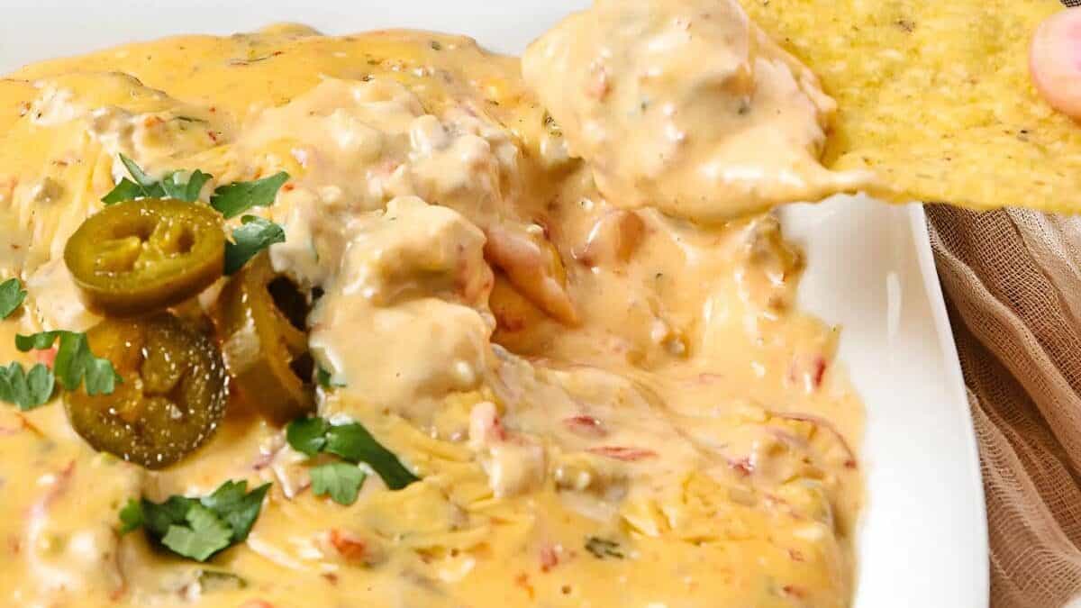A close-up of creamy queso dip with chunks of tomatoes and ground meat, topped with sliced jalapeños and cilantro, with a tortilla chip being dipped into the cheese—perfect for your next round of Dinner Party Recipes.