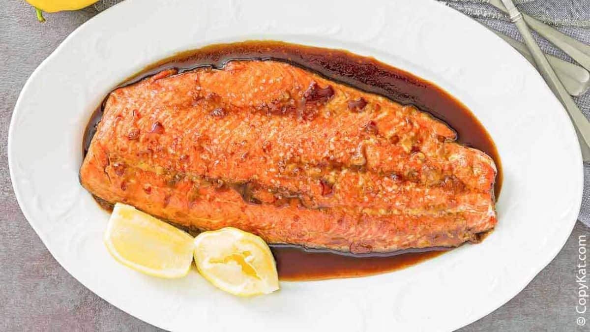 A cooked salmon fillet topped with a brown glaze is served on a white oval plate with two lemon wedges on the side.