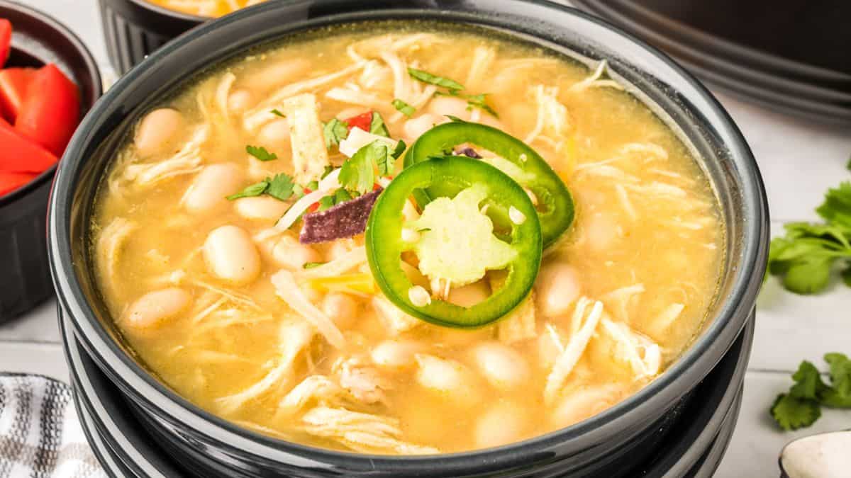 A bowl of white chicken chili topped with sliced jalapeños, cilantro, and tortilla strips, with shredded chicken and white beans visible in a creamy broth. Small bowls of diced tomatoes and cilantro are in the background.