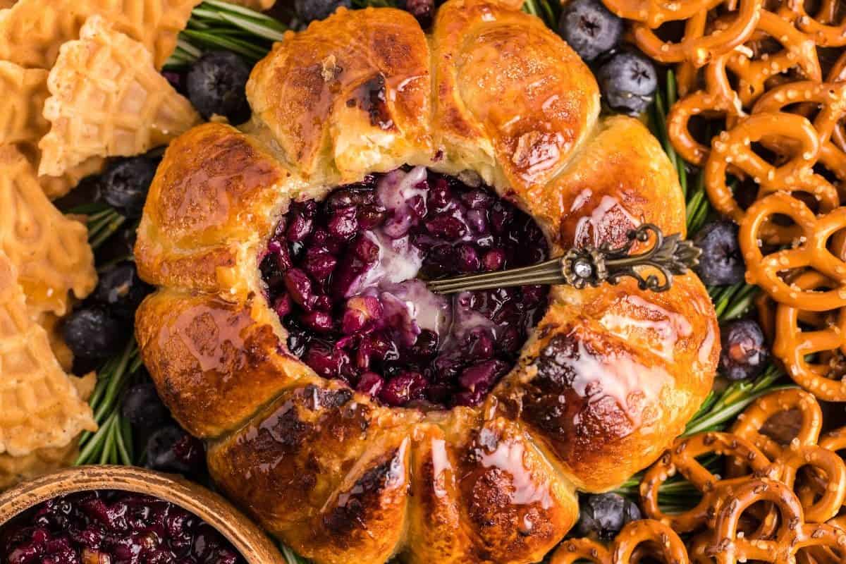 A golden-brown baked pastry filled with dark berry compote is surrounded by pretzels, waffle cookies, blueberries, and rosemary—appetizers too good to share. An ornate spoon rests inside the pastry’s berry-filled center.