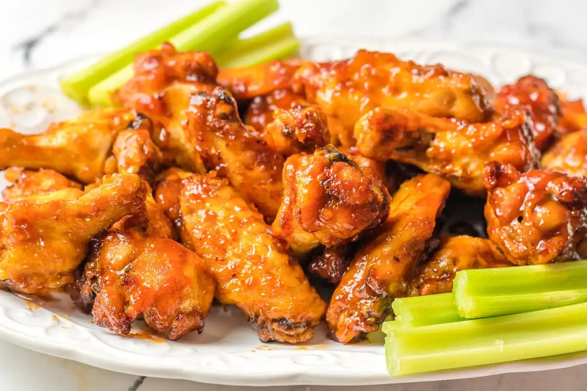 A plate of glazed buffalo chicken wings garnished with sauce, served with fresh celery sticks on the side—appetizers too good to share. The food is arranged on a white platter atop a marble surface.