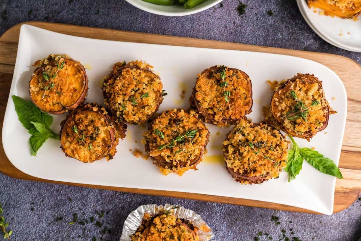 Eight baked stuffed mushrooms topped with golden, crispy breadcrumbs and fresh herbs are arranged on a white platter—perfect alongside fall casseroles that make weeknights feel like Sunday. Basil leaves add a touch of color to the wooden board display.