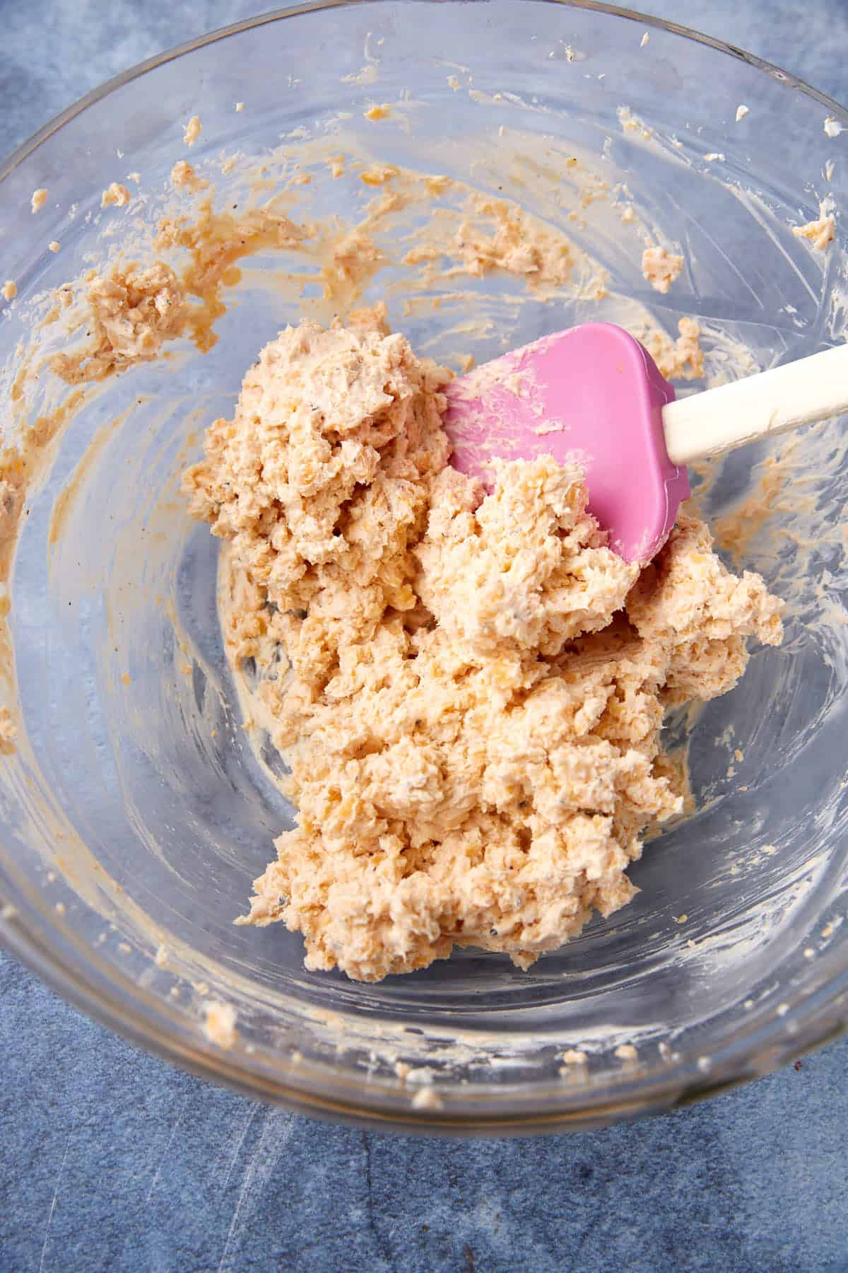 A glass mixing bowl containing a thick, beige dough mixture and a pink silicone spatula, resting on a blue textured surface.