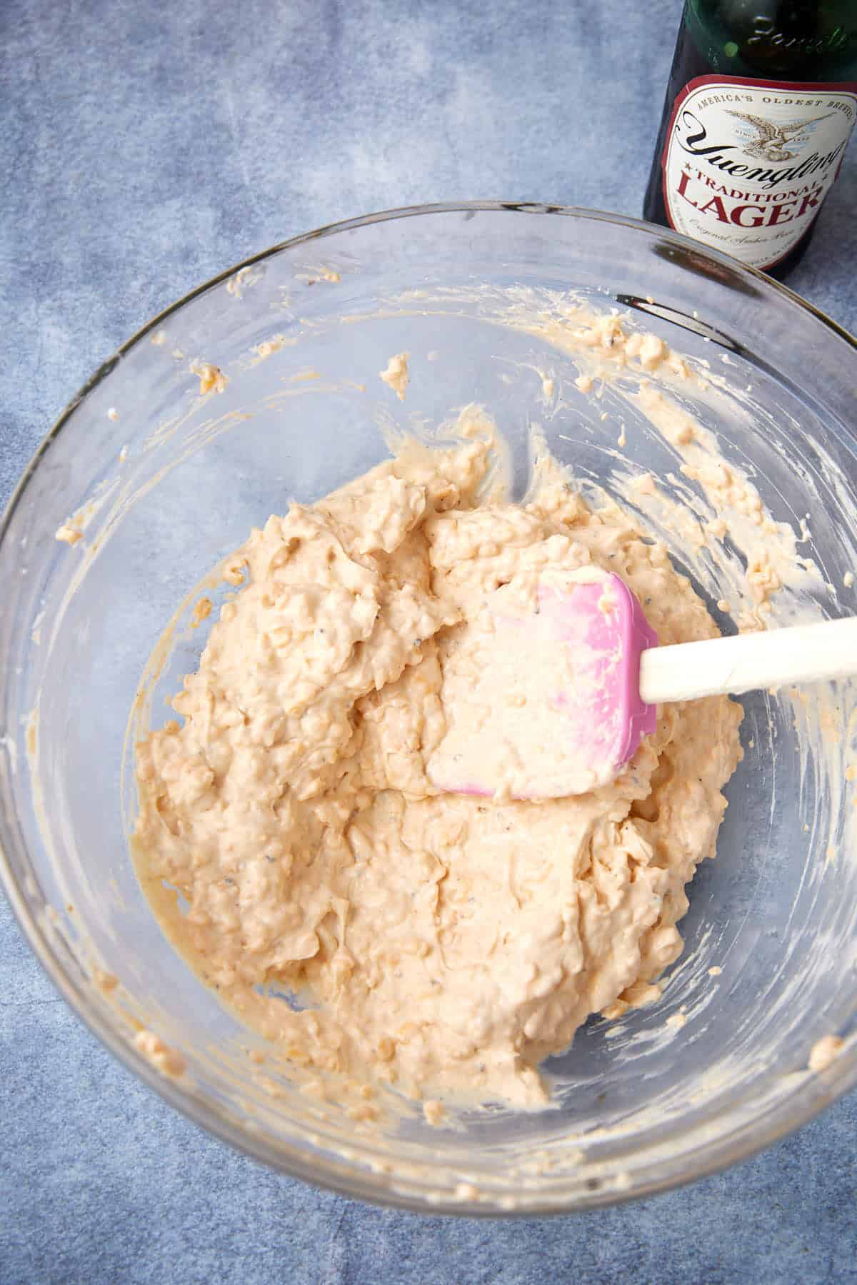 A glass bowl filled with a creamy, chunky mixture being stirred with a pink spatula. A bottle of Yuengling Traditional Lager is partially visible in the top right corner on a blue surface.