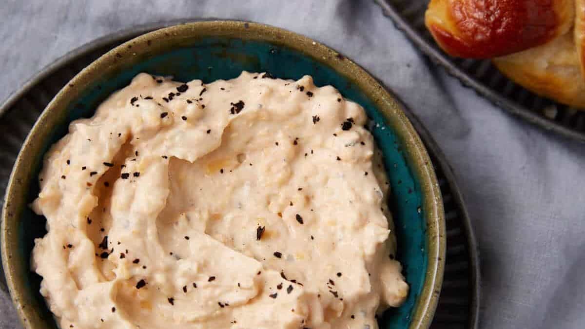 A bowl of creamy, white cheese dip sprinkled with black pepper sits on a plate beside a vintage knife, with soft pretzels on a separate plate nearby, all on a light gray cloth background.