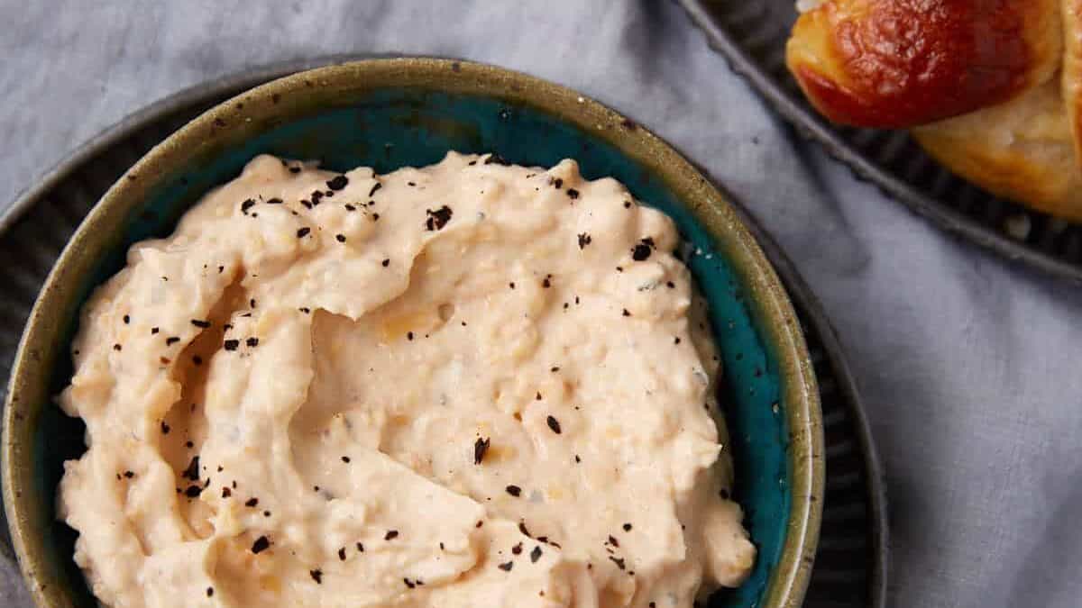 A bowl of creamy, white cheese dip sprinkled with black pepper sits on a plate beside a vintage knife, with soft pretzels on a separate plate nearby, all on a light gray cloth background.