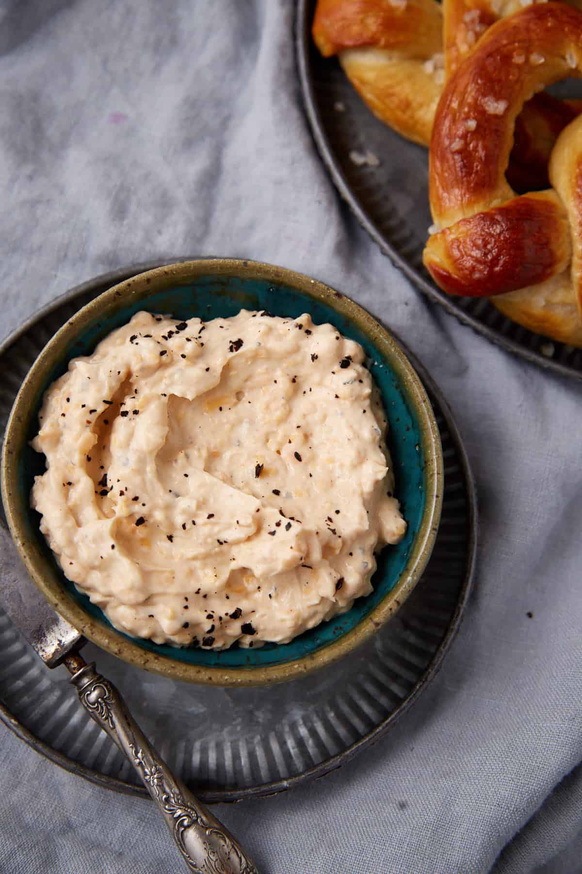 A bowl of creamy, white cheese dip sprinkled with black pepper sits on a plate beside a vintage knife, with soft pretzels on a separate plate nearby, all on a light gray cloth background.