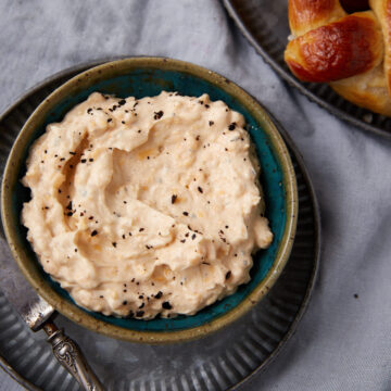 A bowl of creamy spread topped with cracked black pepper, placed on a metal plate with a spoon beside it; part of a pretzel is visible on a separate plate in the corner.