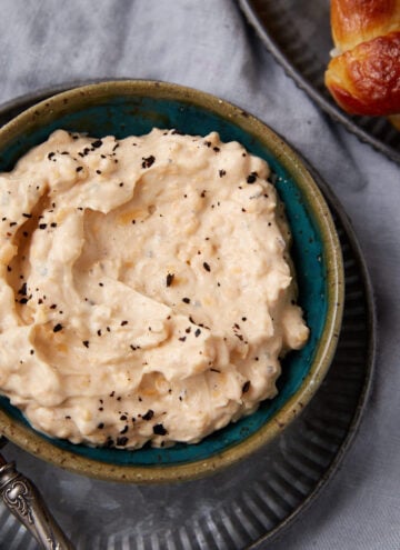 A bowl of creamy spread topped with cracked black pepper, placed on a metal plate with a spoon beside it; part of a pretzel is visible on a separate plate in the corner.