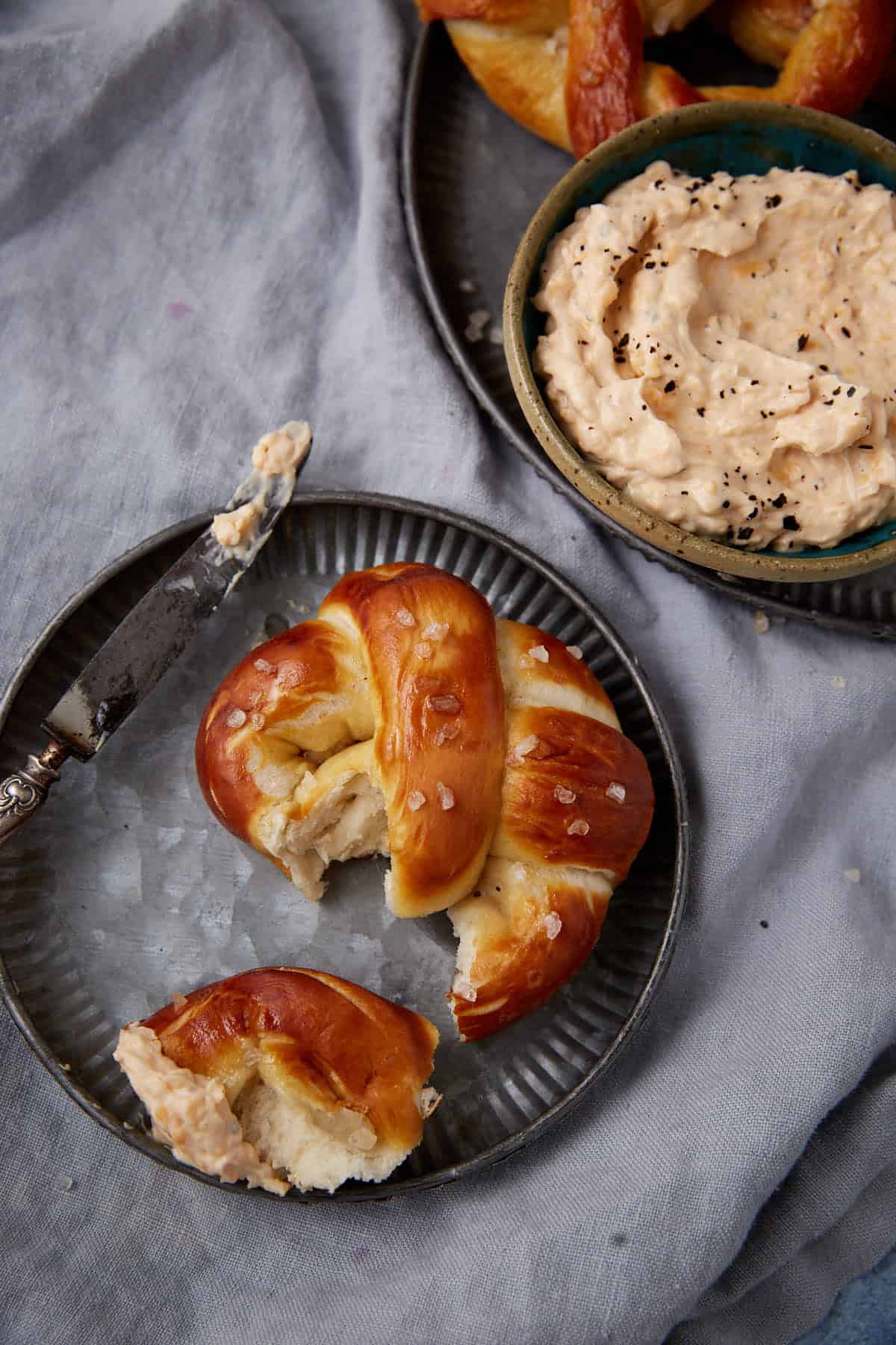 A soft pretzel, partially eaten, sits on a metal plate beside a knife with dip. Nearby, a bowl of creamy dip garnished with black pepper and more pretzels are visible on a gray cloth background.