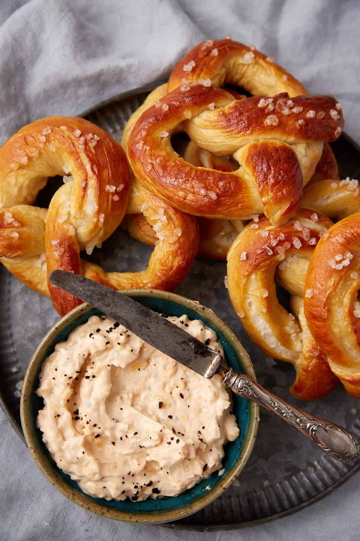 A plate of golden-brown pretzels sprinkled with coarse salt sits next to a bowl of creamy dip topped with cracked black pepper. A vintage knife rests on the bowl, all on a gray cloth background.