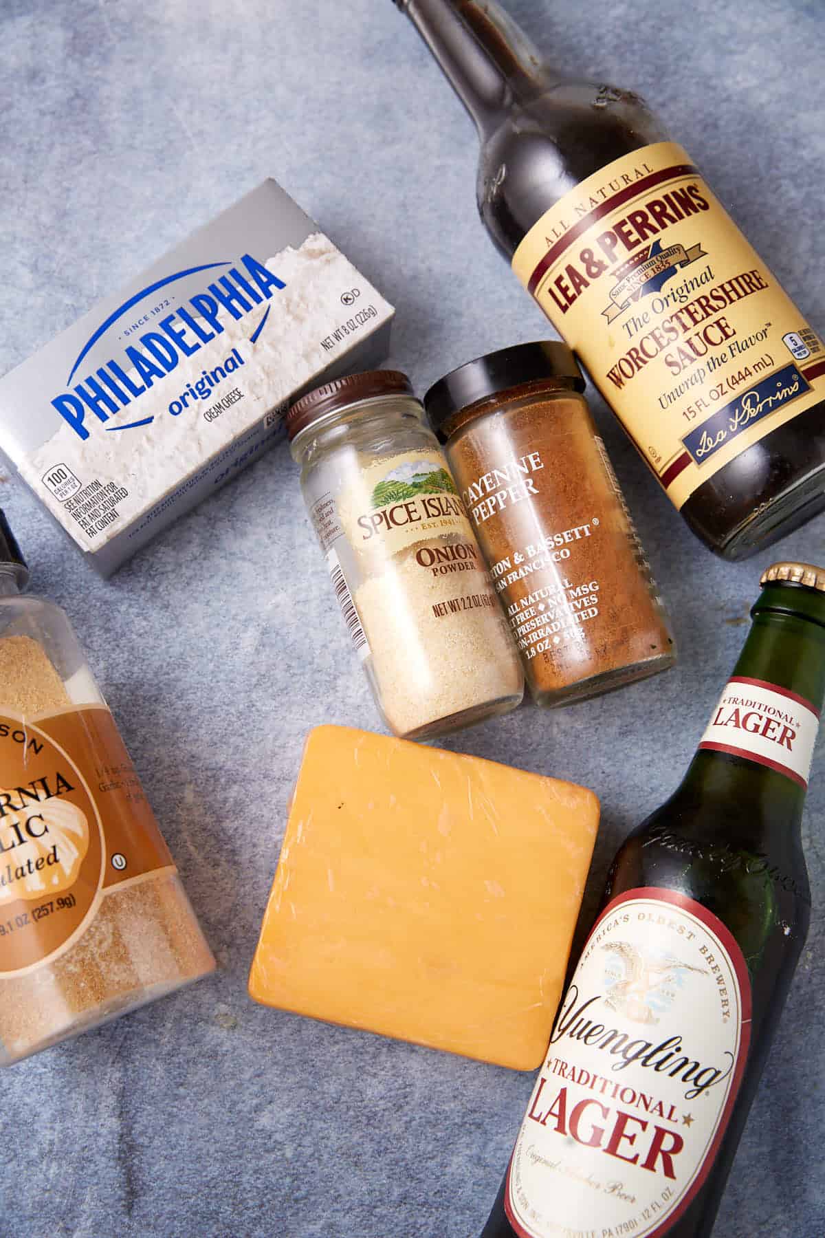 A variety of ingredients on a gray surface, including cream cheese, a block of cheddar, Worcestershire sauce, lager beer, onion powder, cayenne pepper, and brown sugar.