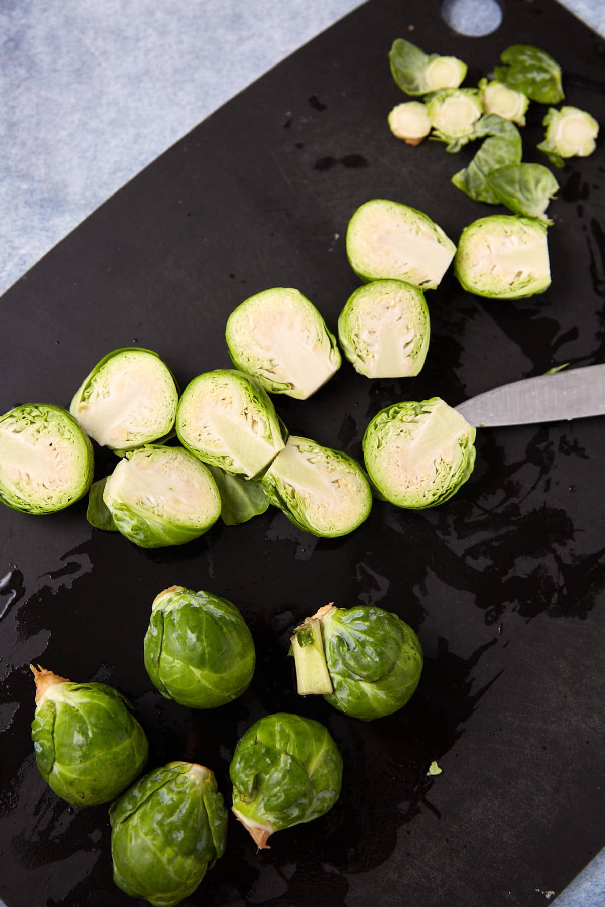 A black cutting board with whole and halved Brussels sprouts, a few trimmed stem ends, and a knife, all on a blue surface.