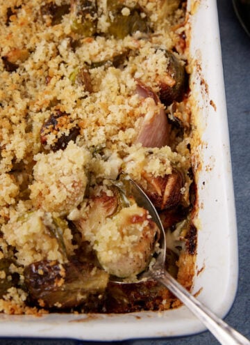 A close-up of a baked casserole dish filled with Brussels sprouts topped with golden, crispy breadcrumbs. A serving spoon is partly inserted, showing the creamy interior.