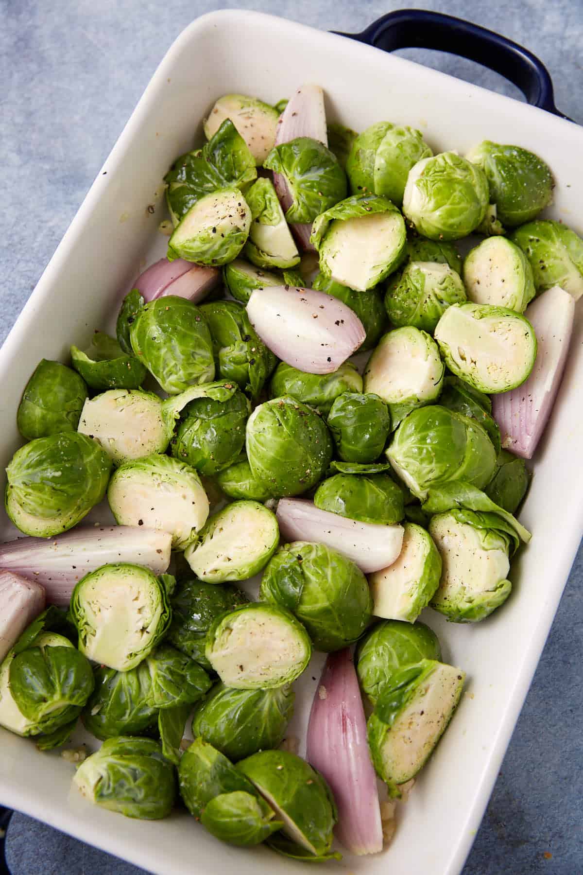 A white baking dish filled with halved Brussels sprouts and quartered shallots, seasoned with black pepper and ready for roasting.