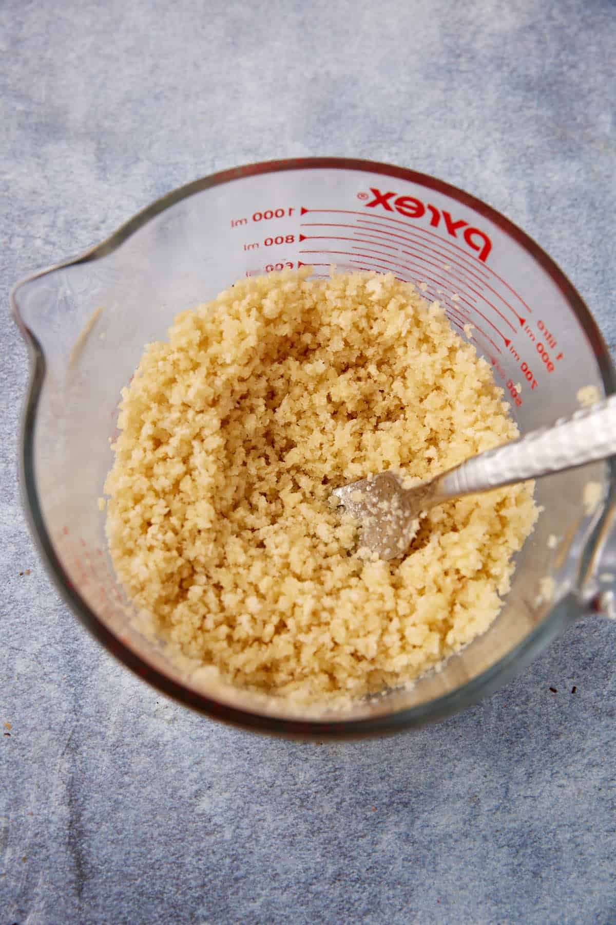 A glass measuring cup containing a crumbly, pale yellow mixture with a metal fork inside, placed on a blue-gray surface.