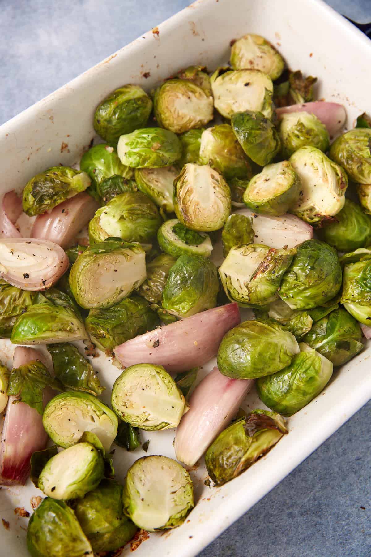 Brussels sprouts and shallots, seasoned and roasted, in a white baking dish on a blue-gray surface.