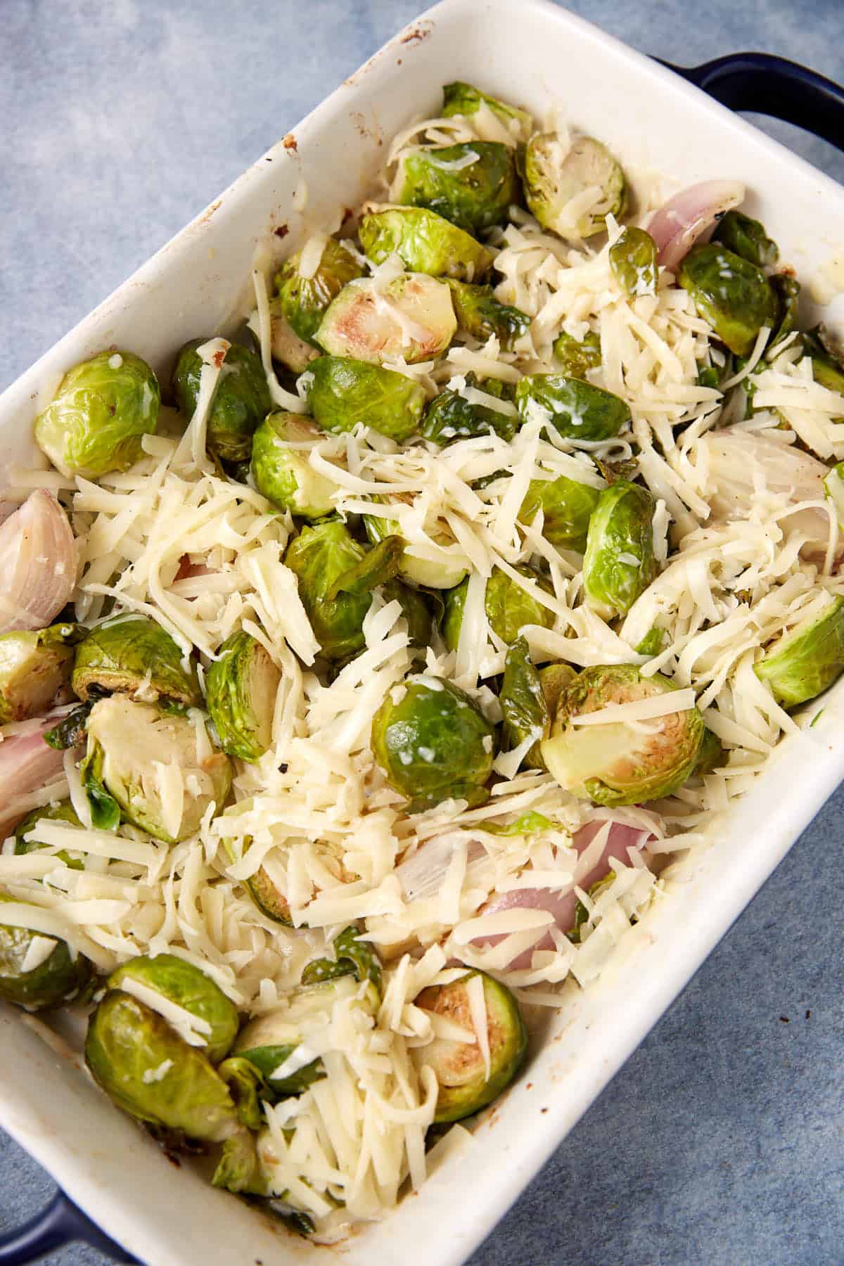 A white baking dish filled with halved Brussels sprouts, sliced shallots, and shredded cheese, ready to be baked. The ingredients are spread evenly in the dish on a gray surface.