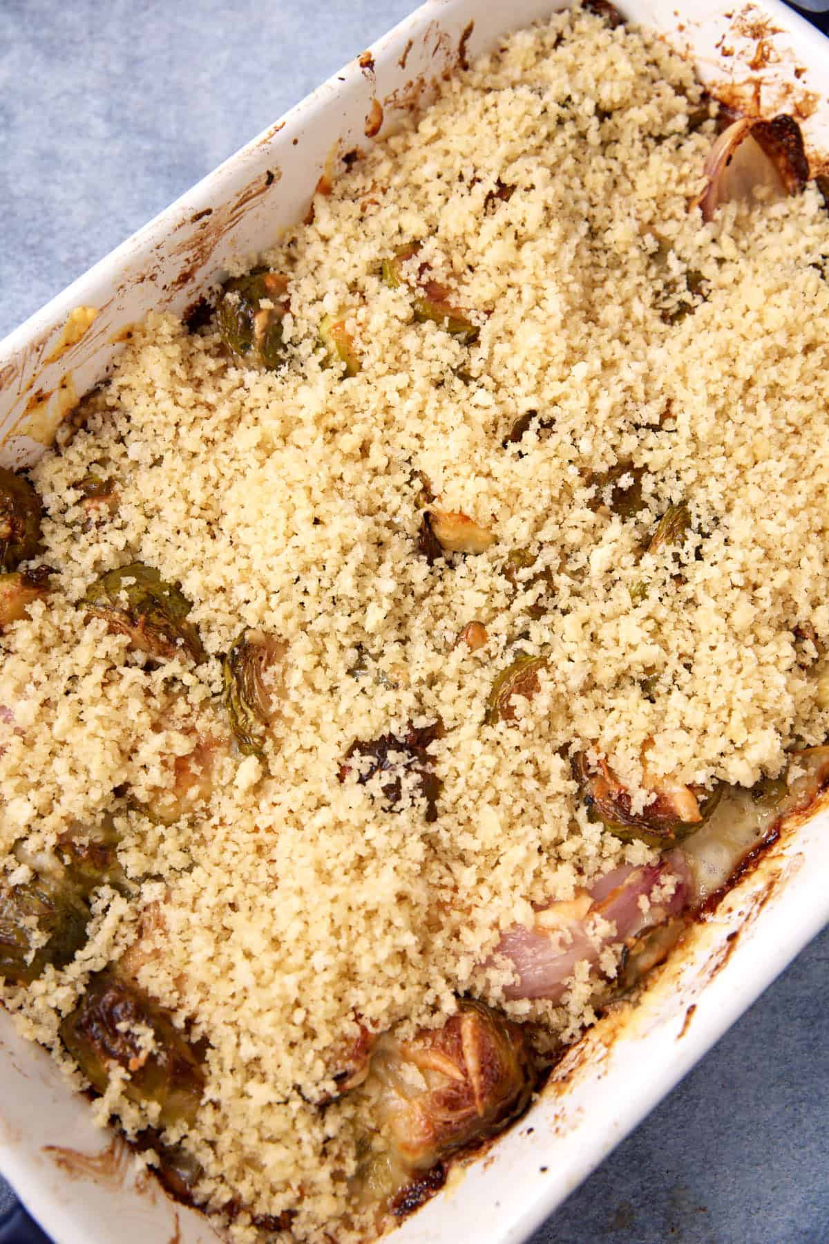 A baked casserole dish filled with roasted vegetables, topped with a golden, crispy breadcrumb layer. The edges of the dish show some browning and bits of caramelized vegetables underneath the crust.