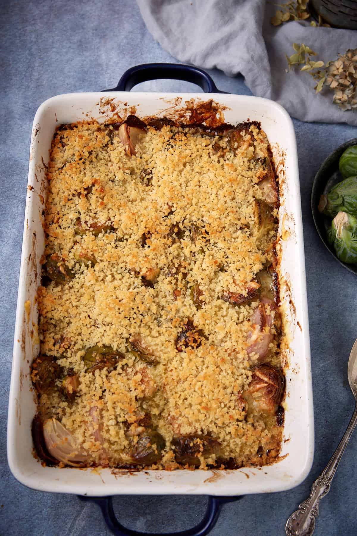 A baked casserole in a white rectangular dish, topped with a golden, crispy breadcrumb crust. Some caramelized vegetables, possibly onions and Brussels sprouts, are visible beneath the topping. The dish rests on a blue surface.