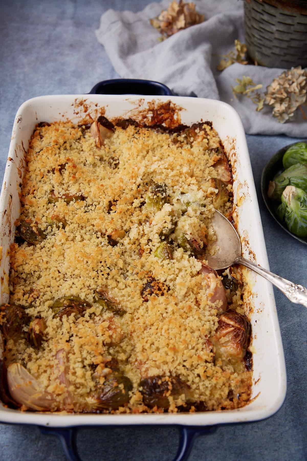 A rectangular baking dish filled with a baked casserole topped with a golden, crispy breadcrumb layer. A serving spoon rests inside the dish. Brussels sprouts and a gray napkin are nearby.