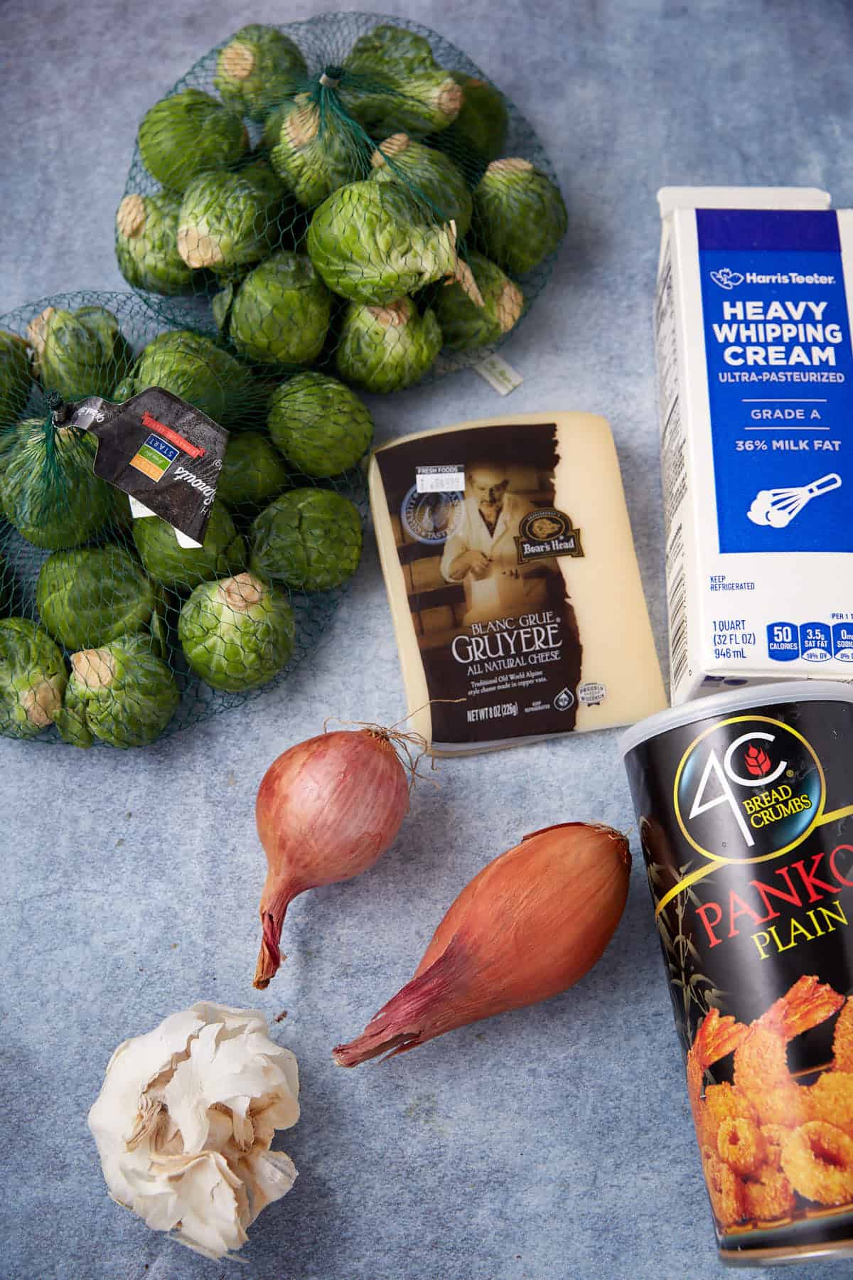 A bag of Brussels sprouts, a wedge of Gruyère cheese, a carton of heavy whipping cream, a container of plain panko breadcrumbs, two shallots, and a head of garlic on a blue surface.