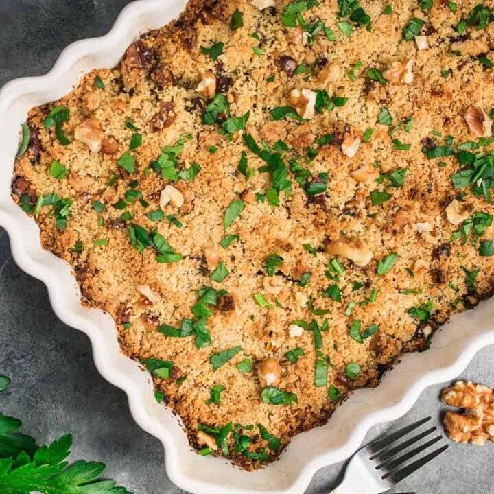 A Butternut Squash Casserole topped with golden breadcrumbs, chopped walnuts, and fresh parsley sits on a gray surface, surrounded by parsley sprigs, whole walnuts, two forks, and a plate.