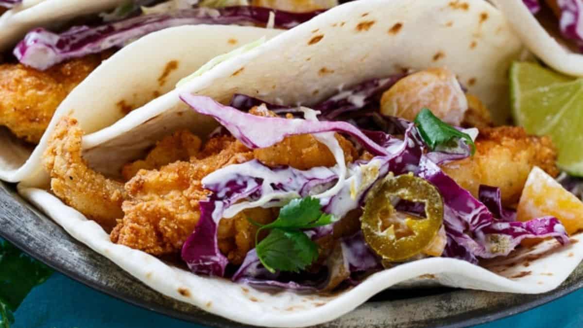 Soft flour tortilla tacos filled with crispy fried fish, shredded purple cabbage slaw, sliced jalapeños, fresh cilantro, and creamy sauce, with a wedge of lime on the side.