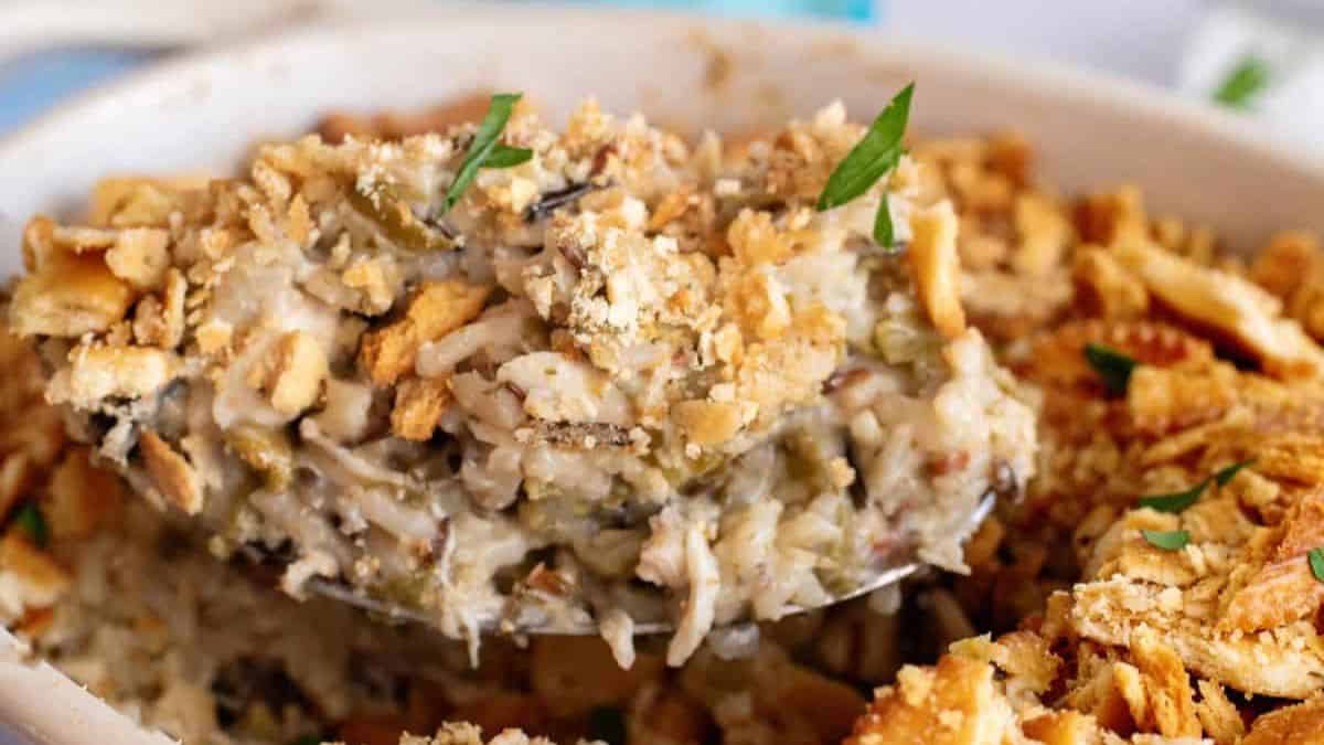 A close-up of a serving spoon lifting a portion of creamy chicken and rice casserole topped with crushed crackers and garnished with chopped parsley.