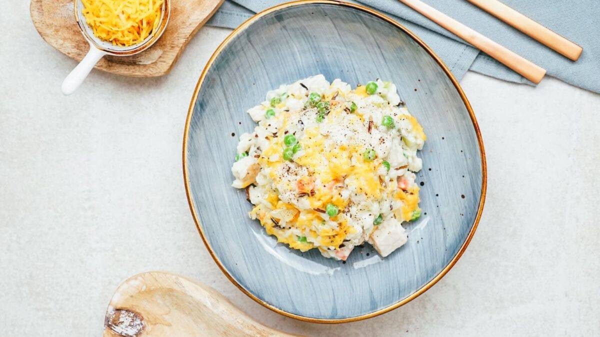 A blue-rimmed plate with a serving of creamy chicken and vegetable casserole topped with melted cheese and black pepper, with a small bowl of shredded cheese and wooden utensils nearby.