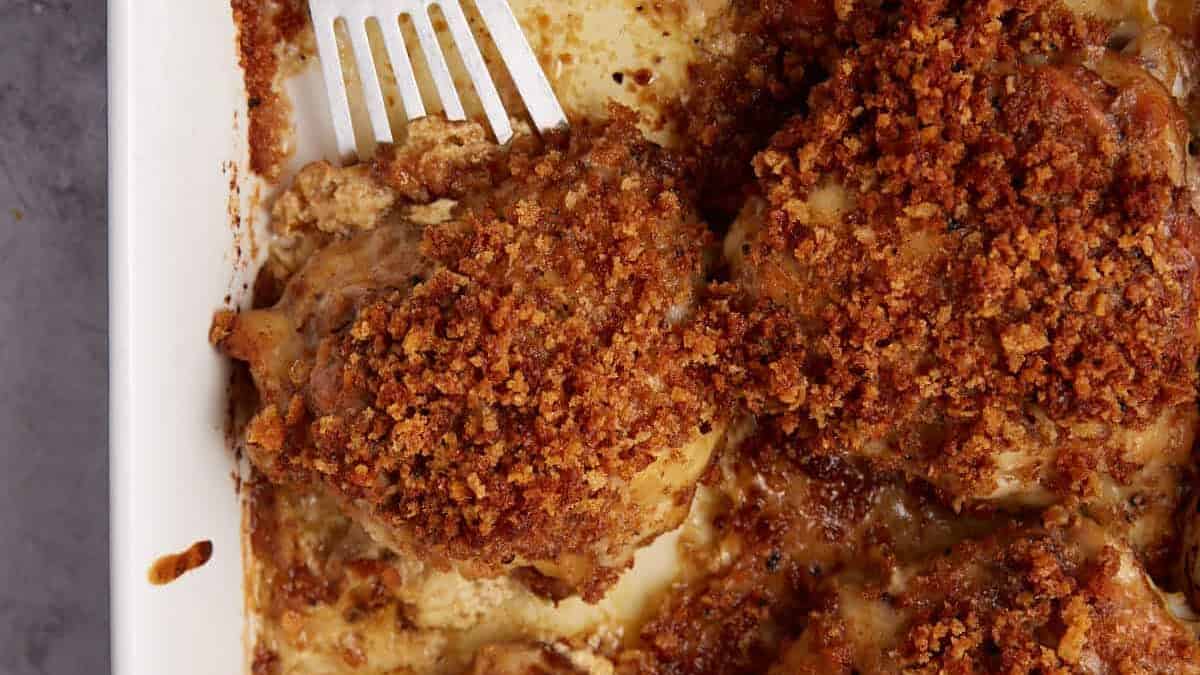 A white baking dish filled with crispy, golden-brown oven baked chicken thighs, some resting in creamy sauce, with a metal spatula lifting one piece.