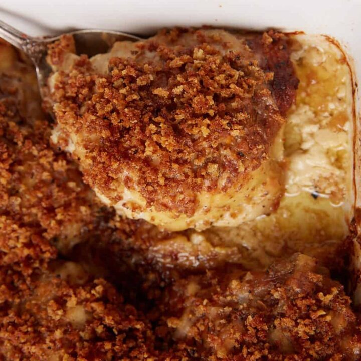 A close-up of a casserole dish filled with golden, crispy oven baked chicken thighs, one piece being served with a spoon. The dish features a browned, crunchy topping and a creamy sauce underneath.