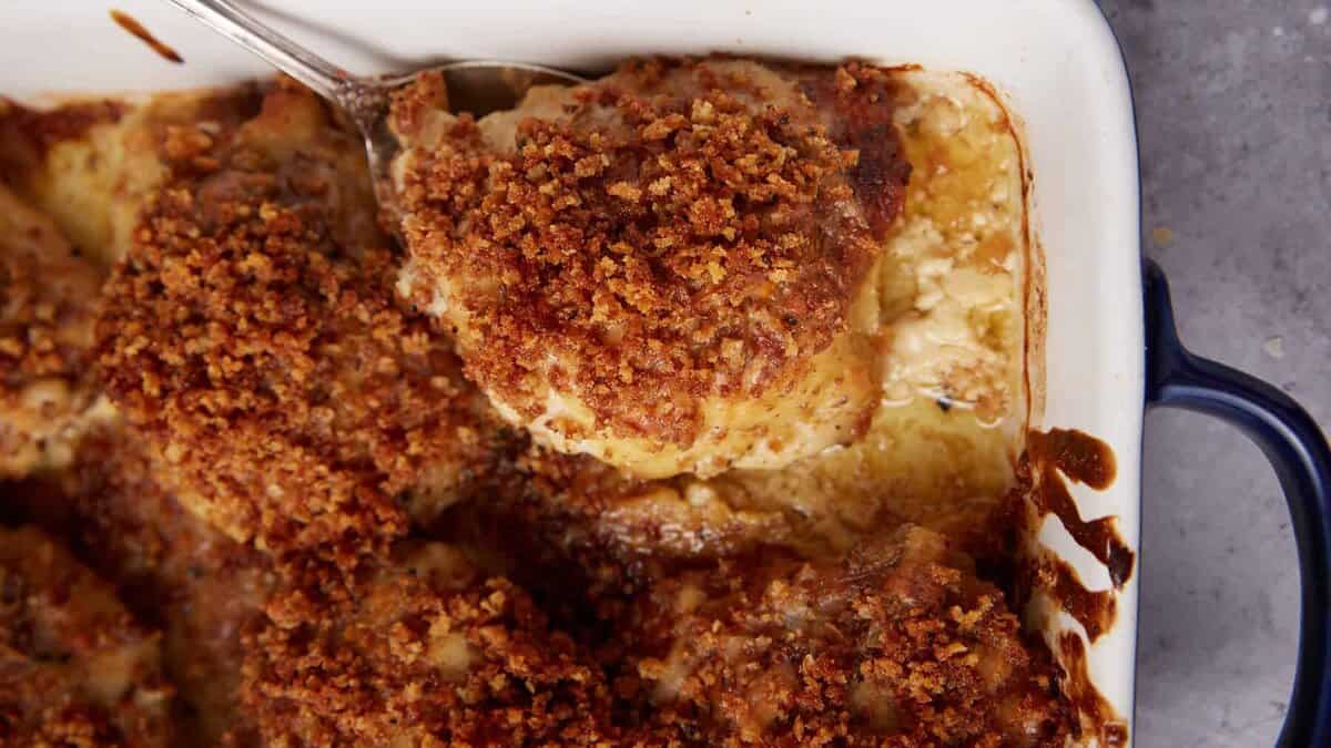 A close-up of a casserole dish filled with golden, crispy oven baked chicken thighs, one piece being served with a spoon. The dish features a browned, crunchy topping and a creamy sauce underneath.
