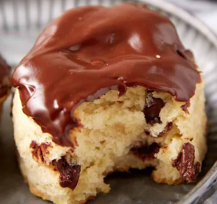 A close-up of a biscuit studded with chocolate chips, partially coated with melted chocolate on top, and with a bite taken out, resting on a metal plate.