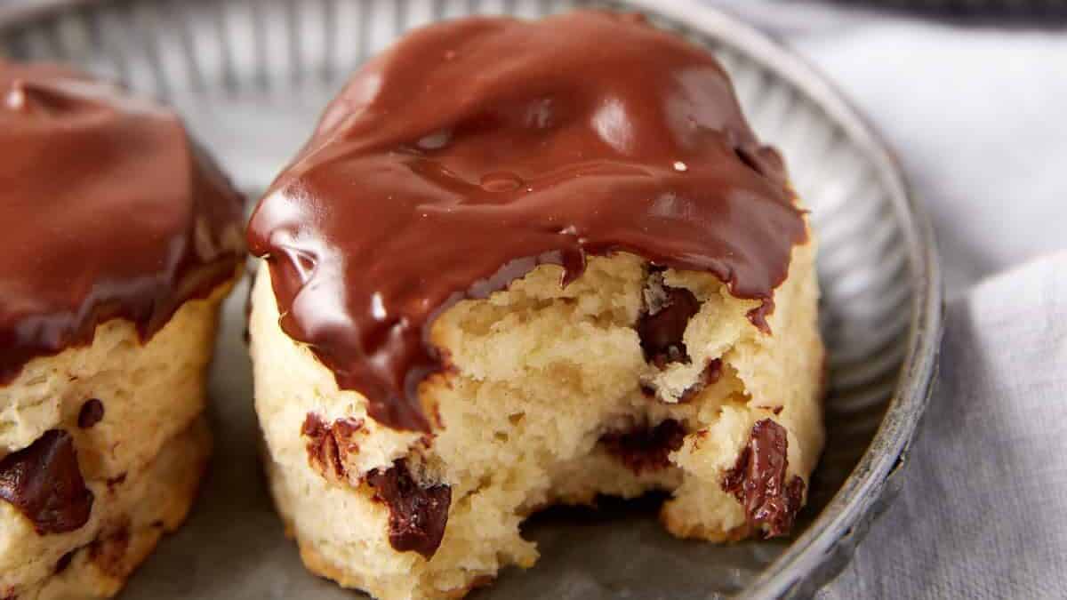 A close-up of a biscuit studded with chocolate chips, partially coated with melted chocolate on top, and with a bite taken out, resting on a metal plate.
