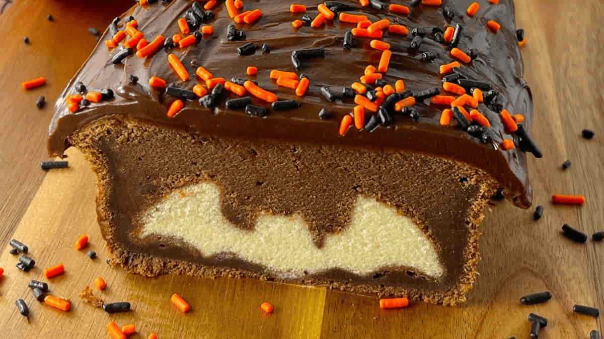 A chocolate loaf cake with chocolate frosting and orange and black sprinkles, sliced to reveal a bat-shaped design in the center of the cake. The loaf sits on a wooden surface with extra sprinkles scattered around.