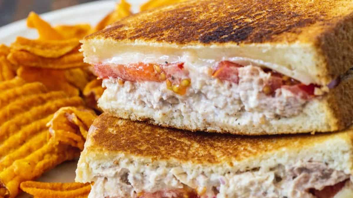 A close-up of a grilled sandwich filled with tuna salad, tomato slices, and cheese, cut in half and stacked on a plate next to a serving of wavy potato chips.