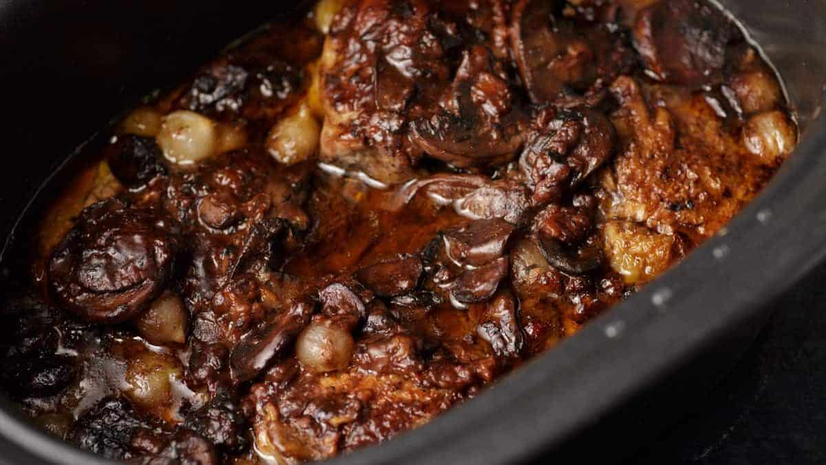 A close-up of a rich, dark beef stew with mushrooms, pearl onions, and a glossy sauce, served in a black pot.