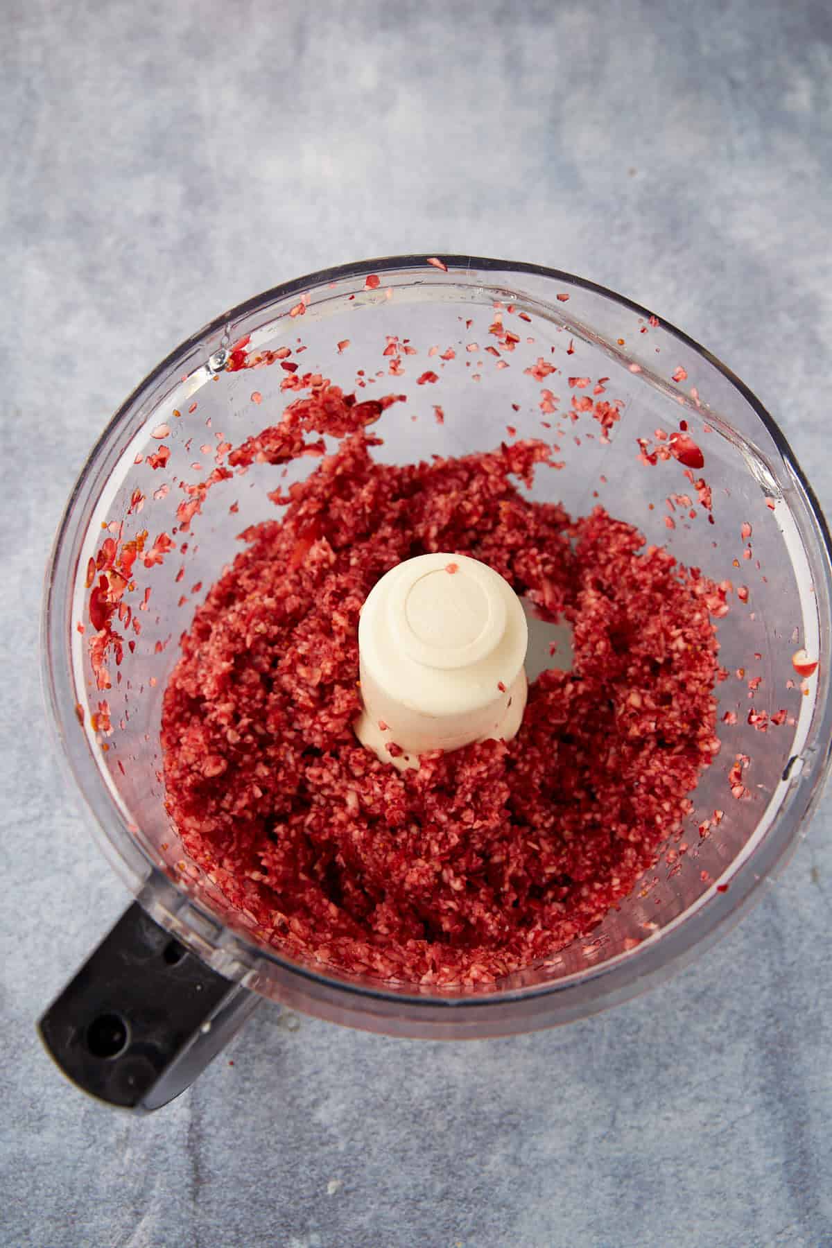 A food processor bowl containing finely chopped or ground red berries and nuts, set on a gray countertop.