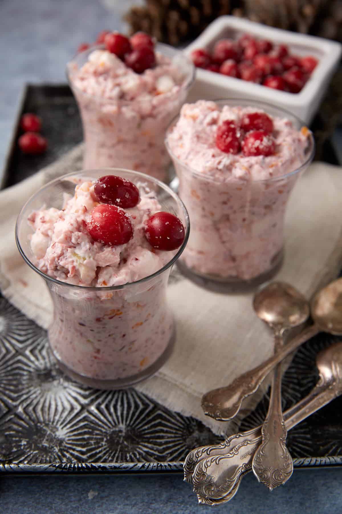 Three glass cups filled with creamy cranberry dessert are placed on a tray with vintage spoons. The dessert is topped with whole cranberries, and more cranberries are visible in a dish in the background.