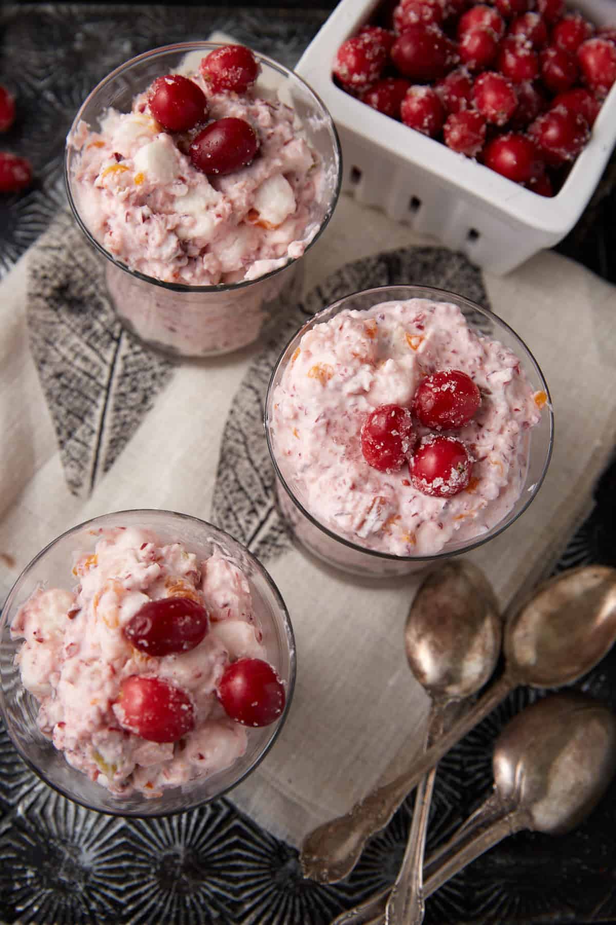 Three glass cups filled with creamy cranberry salad topped with whole cranberries are arranged on a cloth napkin. Nearby, there is a white dish of cranberries and three vintage silver spoons.