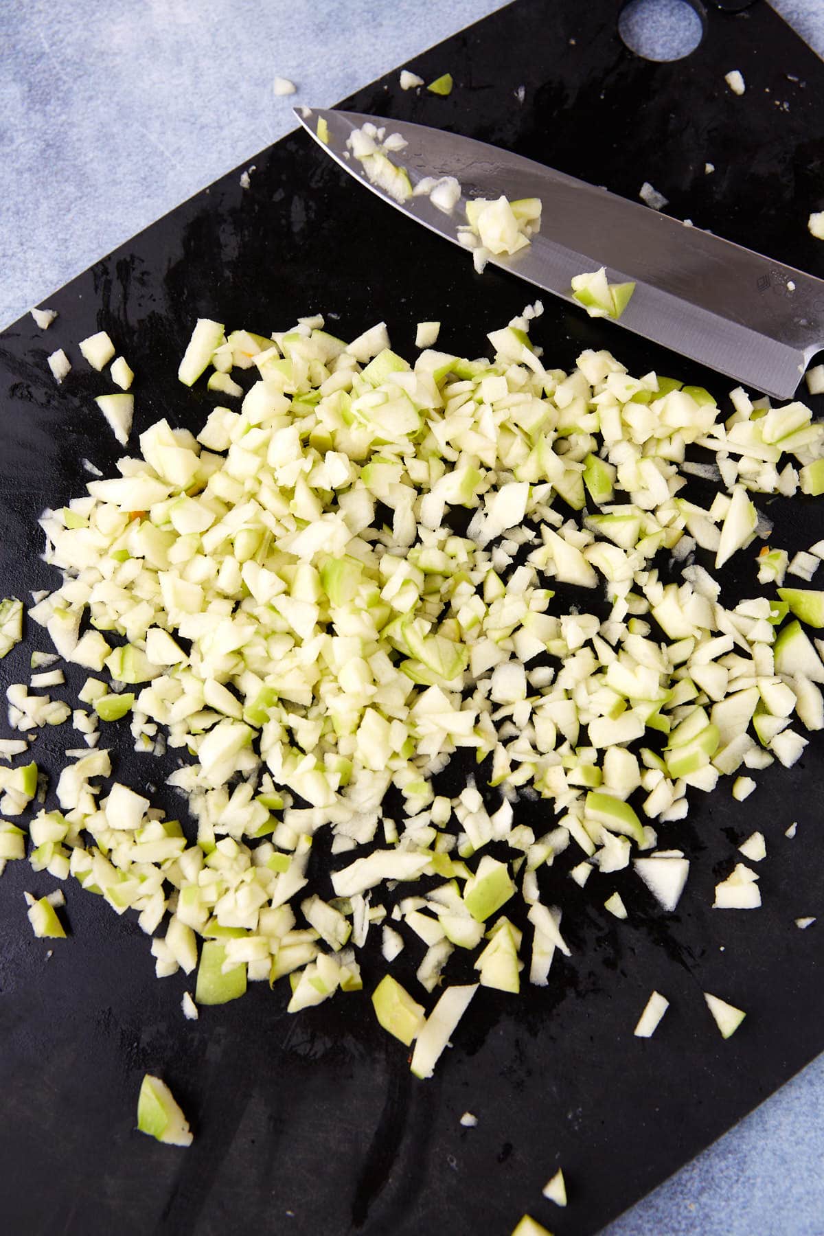 A large kitchen knife and a pile of finely chopped green apple pieces are on a black cutting board, set on a light blue surface.
