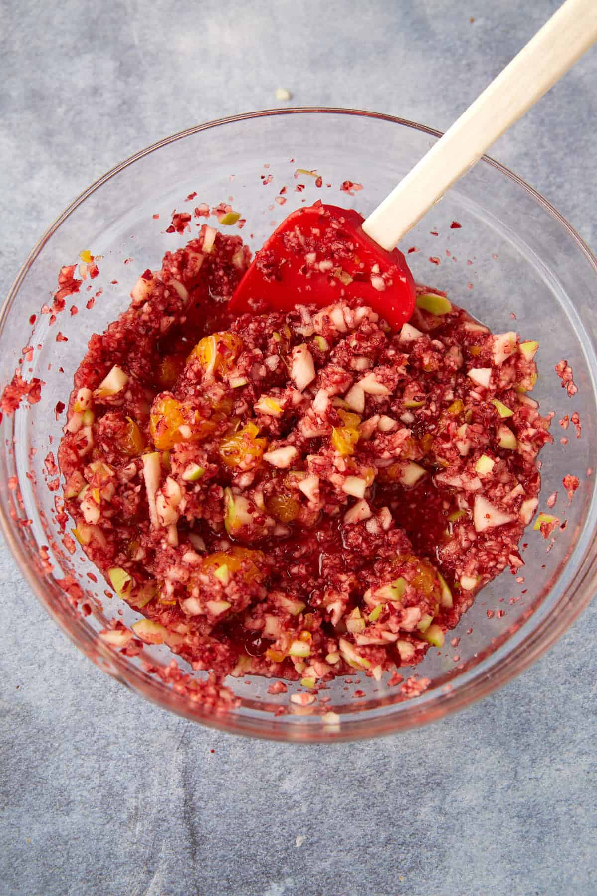 A glass bowl filled with a chopped cranberry mixture, including pieces of apple and orange, being mixed with a red spatula on a gray surface.