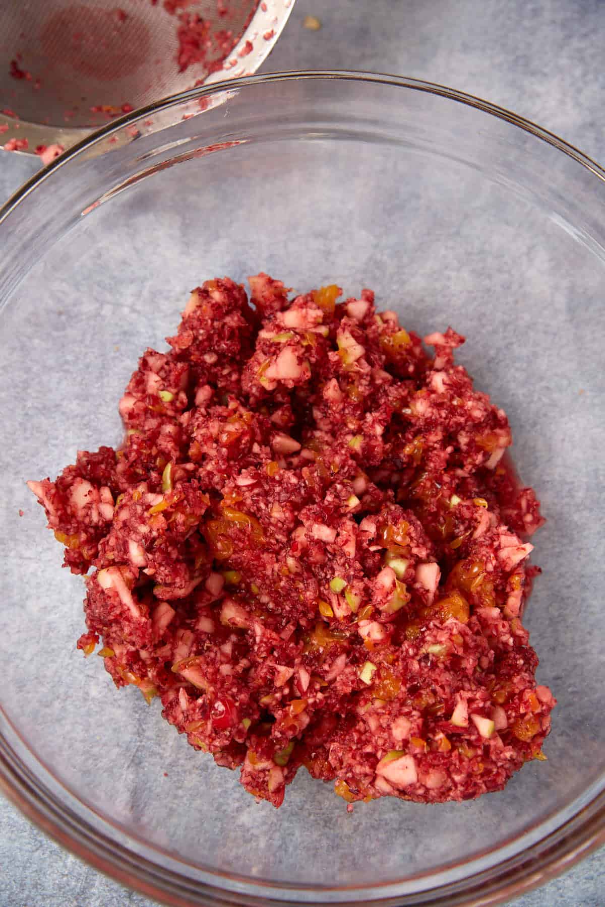 A clear glass bowl containing a chunky mixture of finely chopped red and orange ingredients, likely a fruit or vegetable relish, sits on a gray surface.