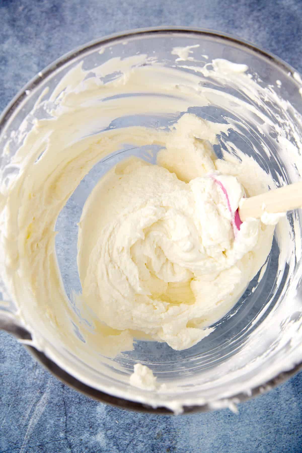 A glass mixing bowl containing thick, creamy batter being stirred with a spatula on a blue countertop.
